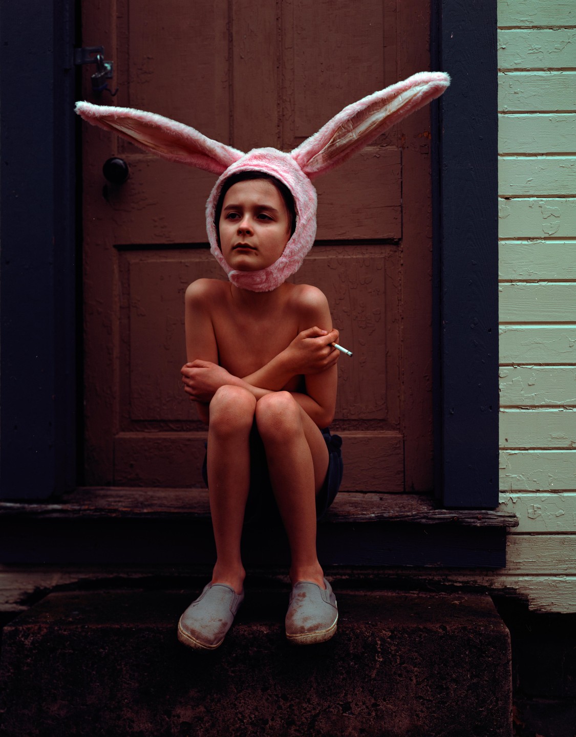 A child wearing a pink bunny hat sits on a step with a cigarette in hand.