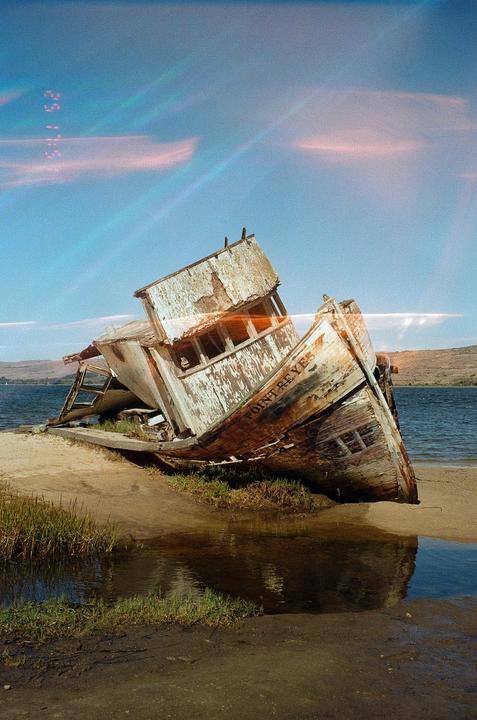 A derelict boat named Point Reyes is beached on a sandy shore next to a body of water under a blue sky.