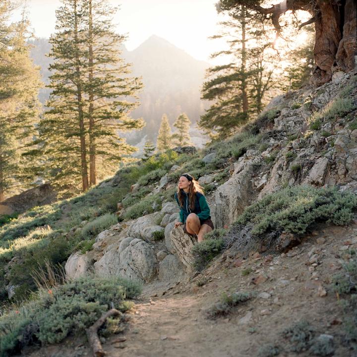 A woman crouches on a rocky mountain path surrounded by pine trees and shrubs, with a hazy mountain in the background.