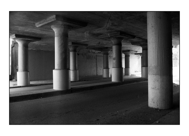 A series of concrete pillars support an overpass above a road, with light visible at the end of the tunnel.