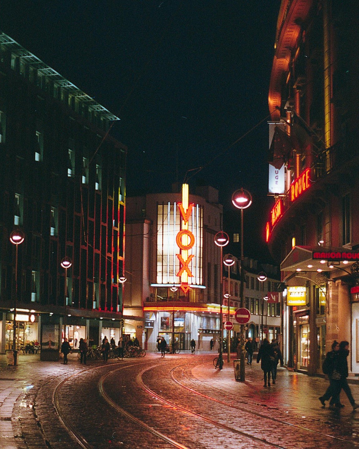 A street with tram tracks curves past buildings with neon signs at night.
