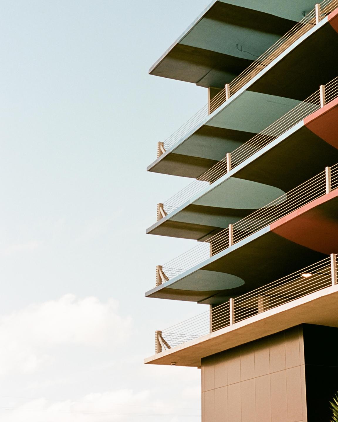 A modern building with multiple levels of balconies and colorful undersides is shown against a clear sky.