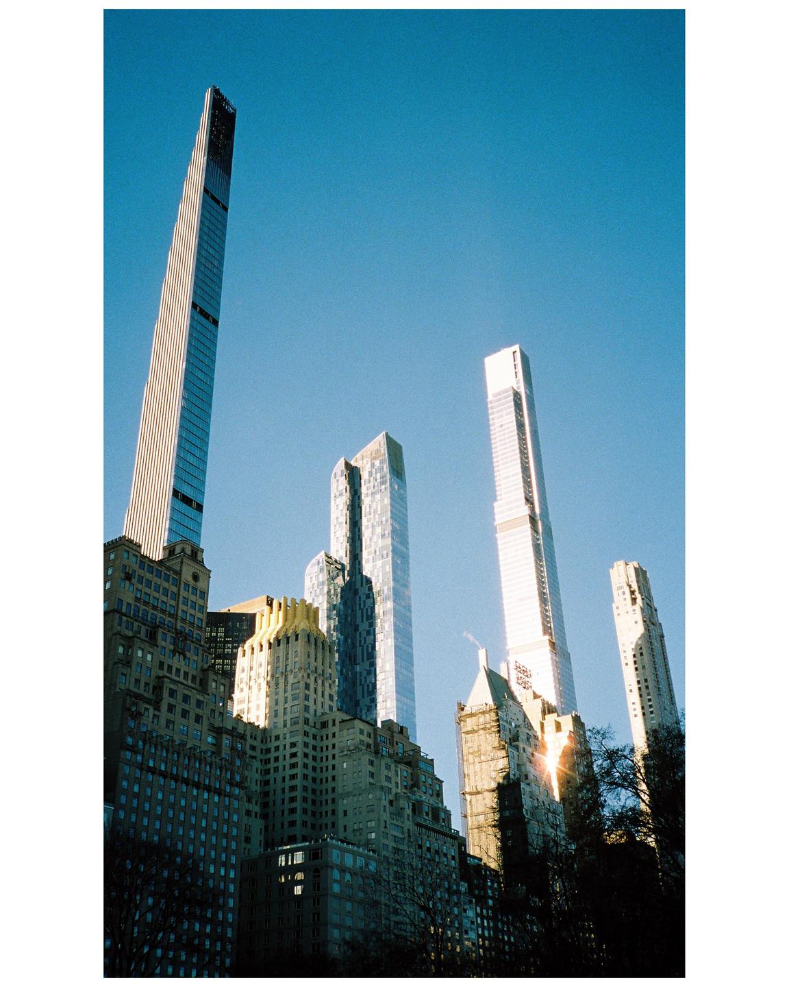 A low-angle view shows several skyscrapers towering over older buildings and trees against a clear blue sky.