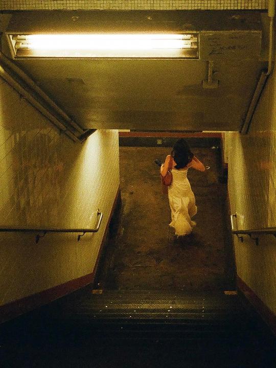 A woman in a light-colored dress walks down a subway staircase illuminated by a fluorescent light.