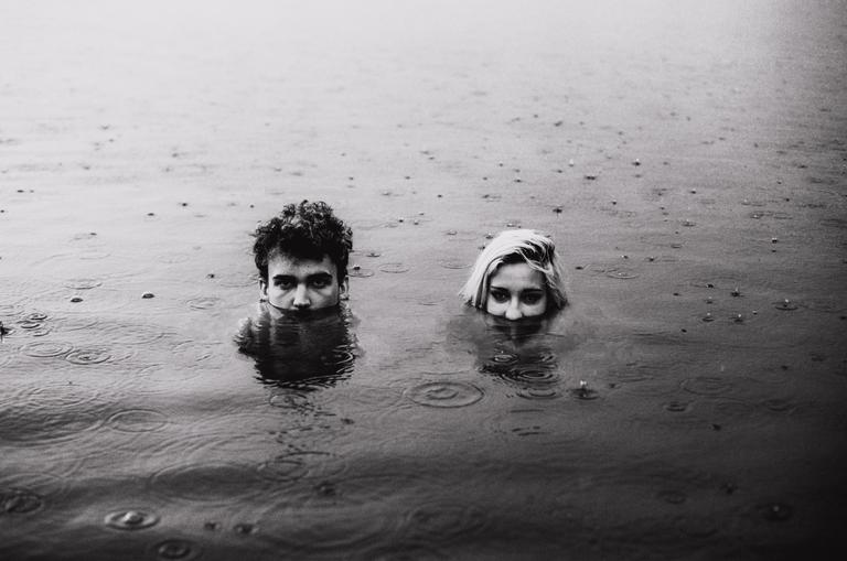 A man and a woman are submerged in water with only their heads visible, as rain falls around them.
