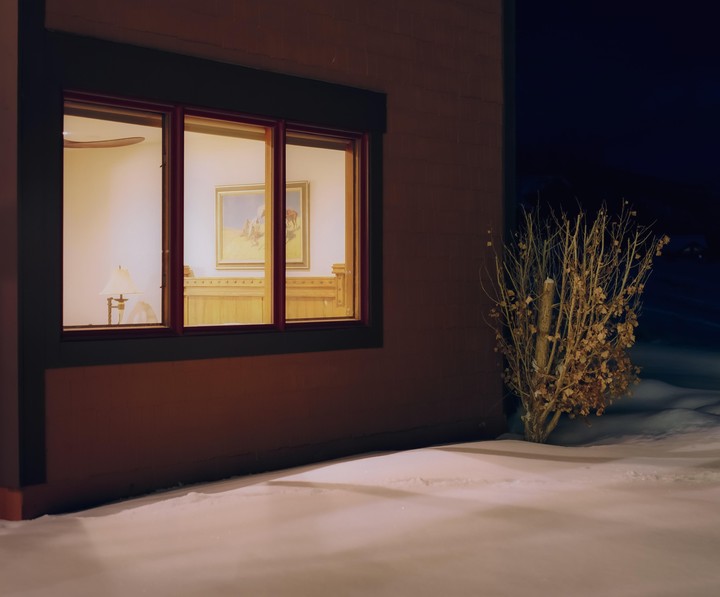 A warm light emanates from a window of a building at night, illuminating a snowy ground and a dry bush outside.