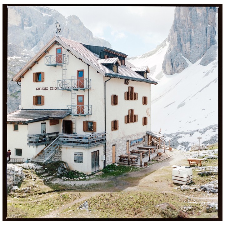 A large white building with brown shutters and balconies stands in a mountainous, snowy landscape with some green grass in the foreground.