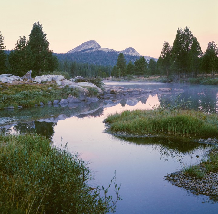 A river with mist on its surface reflects mountains and trees under a pale sky.