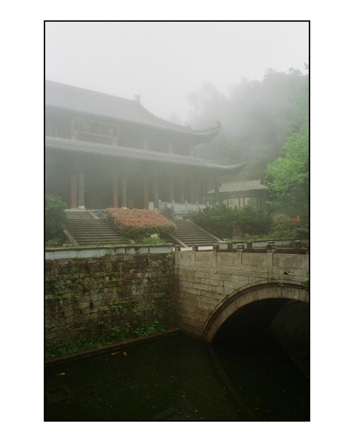 A traditional temple with a curved roof stands behind a stone bridge over a canal, all shrouded in mist.