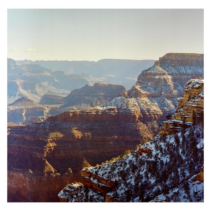 A vast canyon landscape with snow-dusted rock formations is illuminated by sunlight.