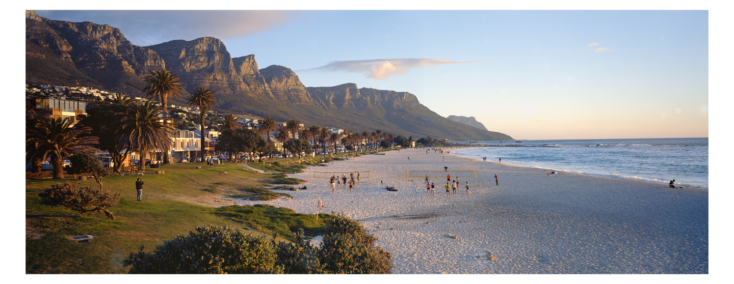 A wide panoramic view shows a beach with people playing volleyball, palm trees, buildings, and mountains under a clear sky.
