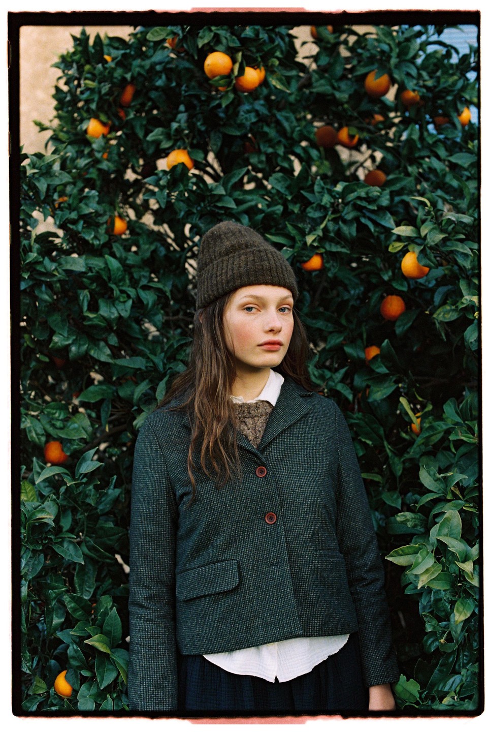 A young woman wearing a beanie and jacket stands in front of an orange tree.