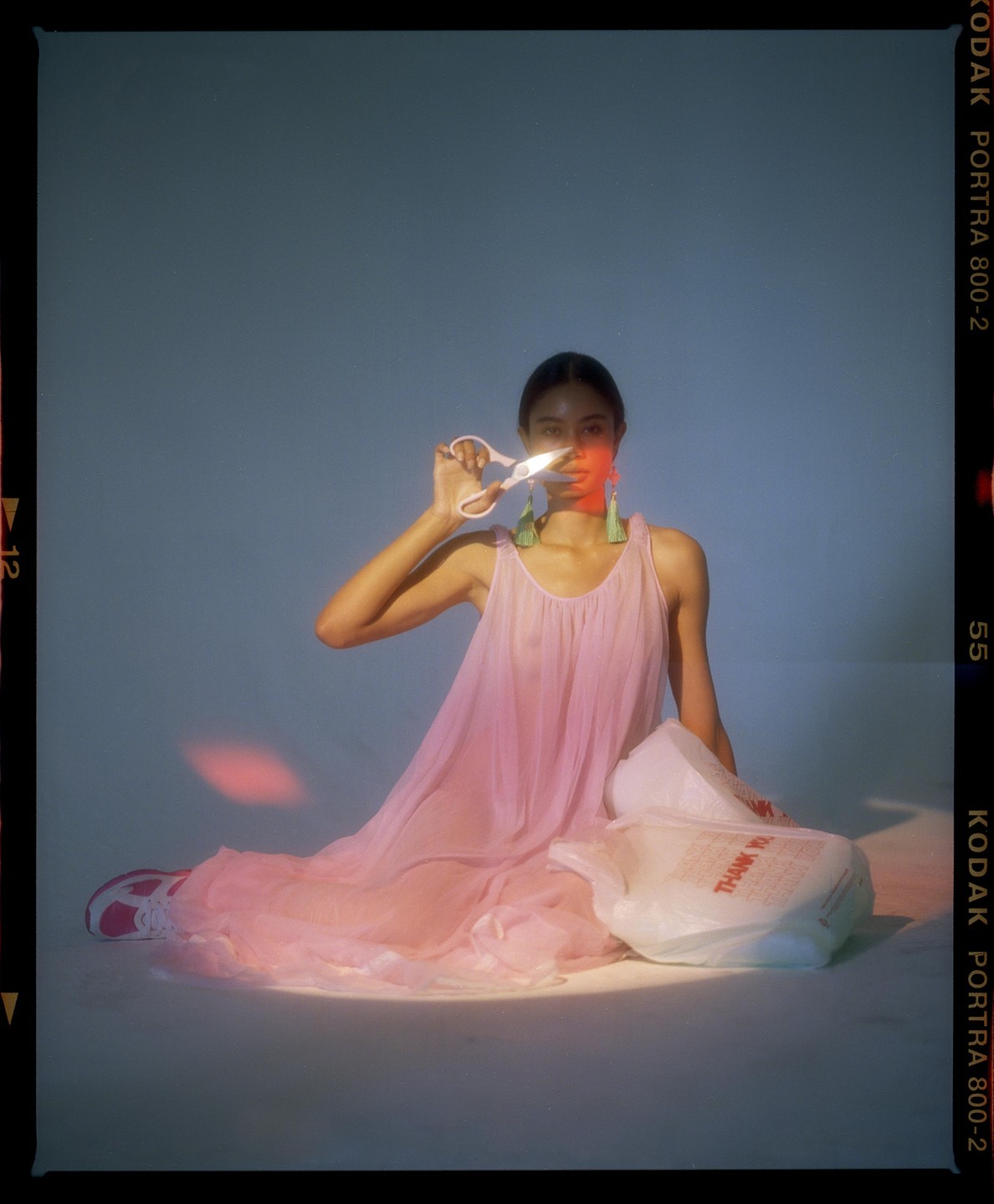 A woman in a pink dress holds scissors in front of her face while sitting on the floor next to two plastic bags.