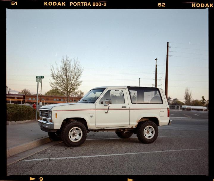 A white Ford Bronco is parked on a street next to a curb with a street sign in the background.