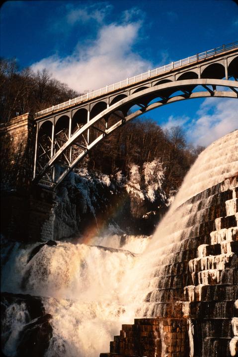 A rainbow appears in the mist of a waterfall next to a bridge and a stepped dam.