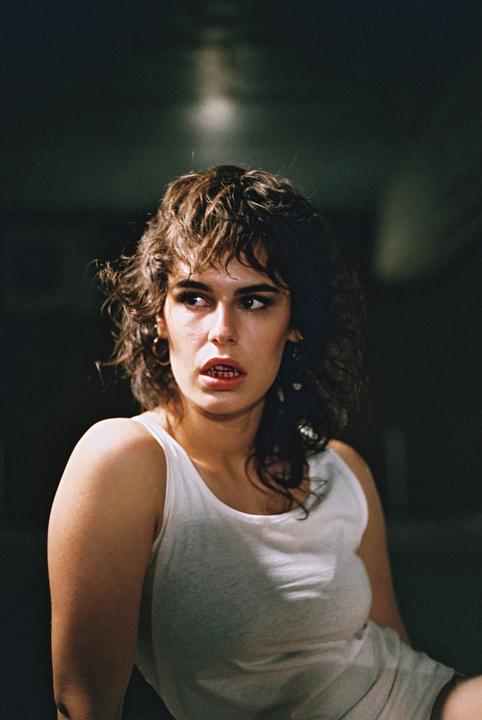 A woman with curly hair and a white tank top looks to the side with her mouth slightly open.