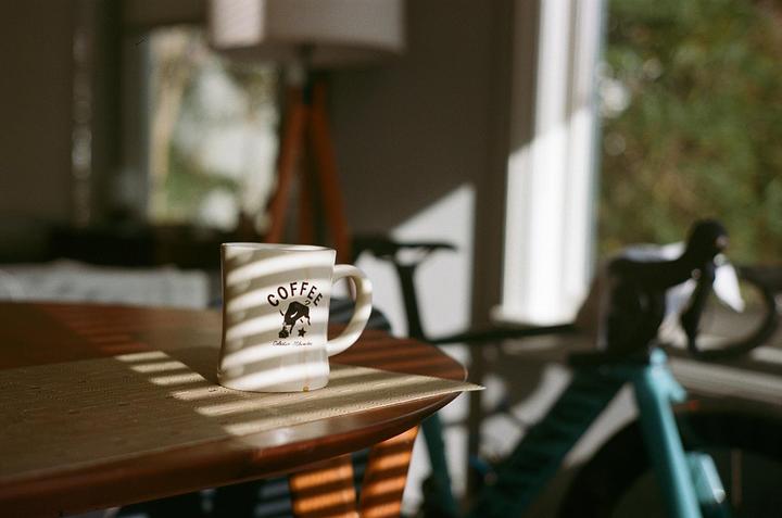 A white coffee mug with a black logo sits on a wooden table with striped shadows.