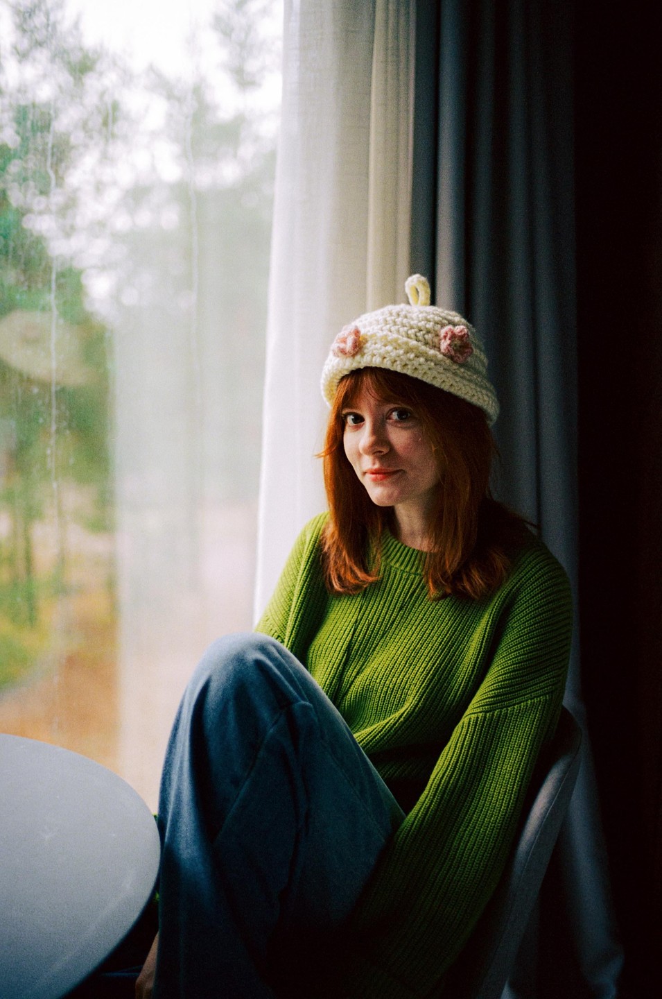 A woman with red hair and a green sweater sits by a window, wearing a white knitted hat with pink flowers.