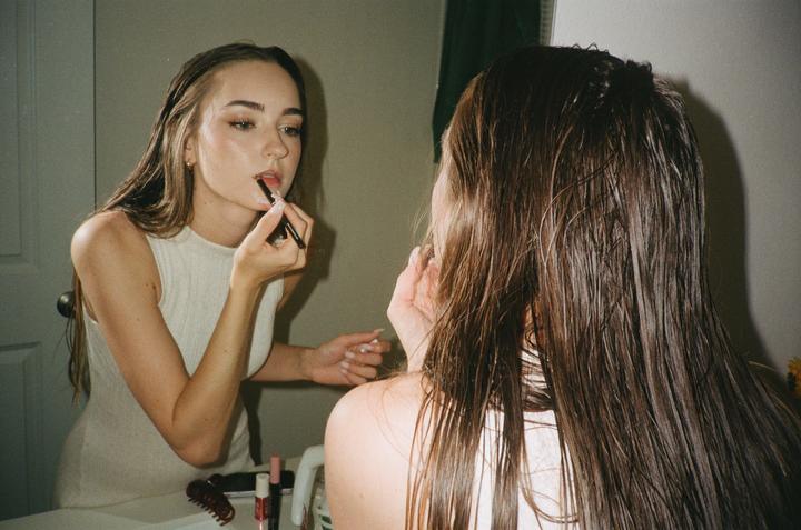 A woman applies lipstick while looking at her reflection in a mirror.