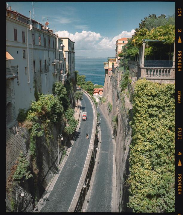 A narrow road leads down to the sea, flanked by buildings and steep, overgrown cliffs.
