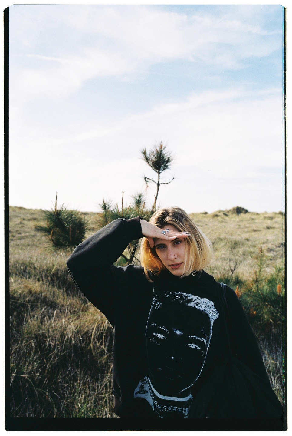A woman shields her eyes from the sun in a field with sparse trees.