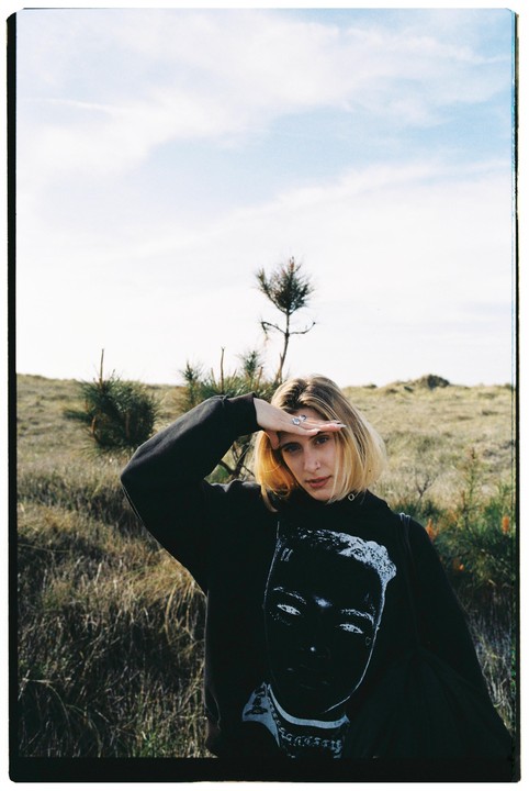 A woman shields her eyes from the sun in a field with sparse trees.