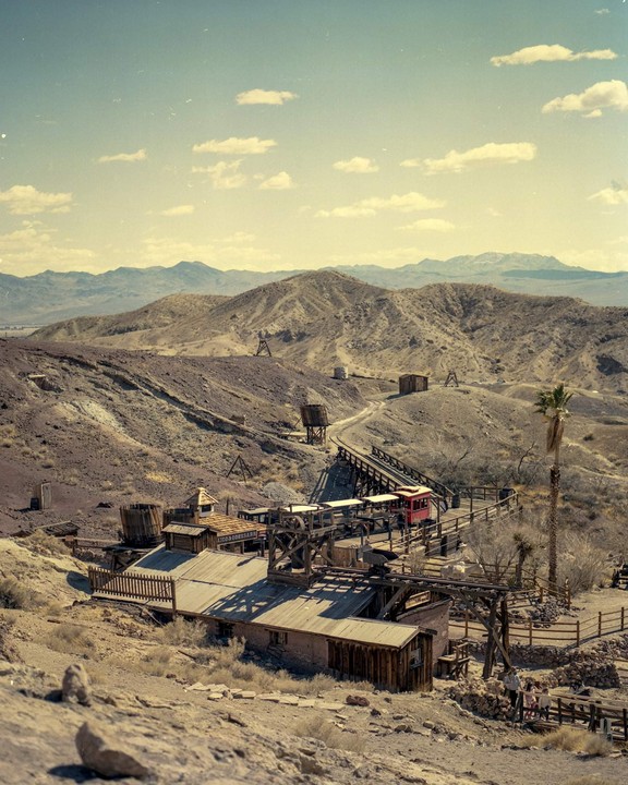 A red mining cart travels along a track above a collection of buildings in a desert landscape.