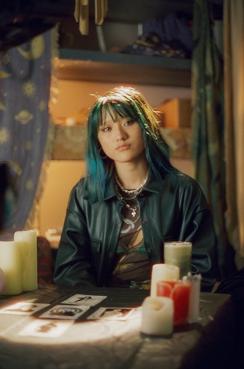A woman with blue hair sits at a table with tarot cards and candles.