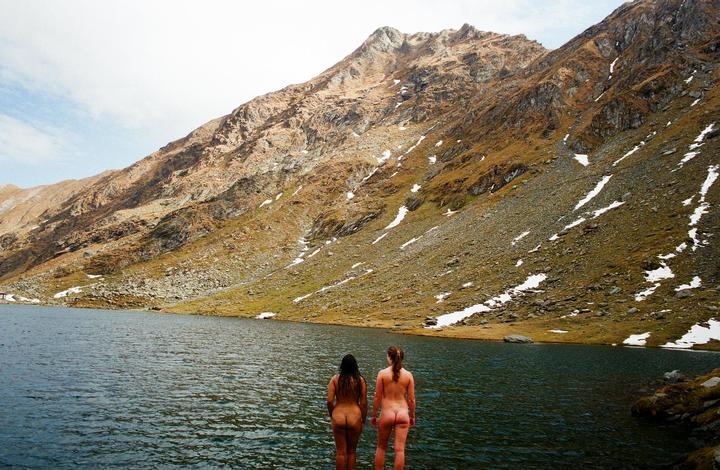 Two women stand naked at the edge of a mountain lake with a rocky, snow-dusted mountain in the background.