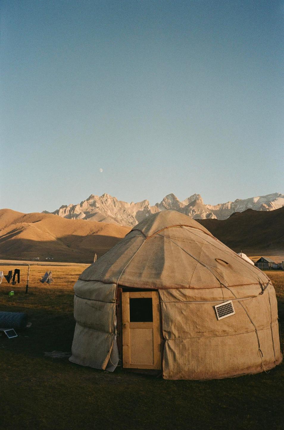 A yurt stands in a grassy field with mountains and a moon in the background.