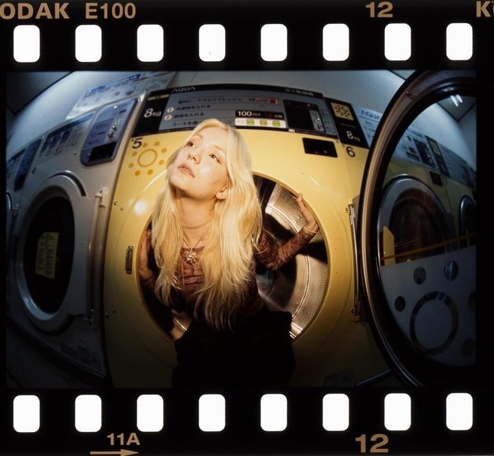 A woman with long blonde hair looks up while sitting inside a yellow washing machine in a laundromat.