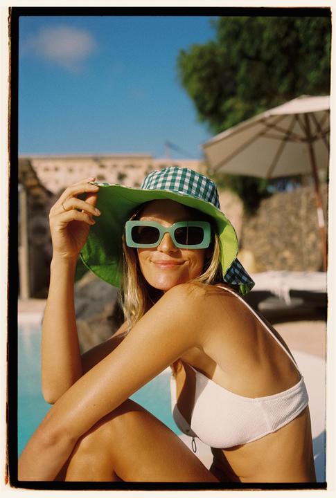 A woman wearing a green hat and sunglasses smiles while sitting by a pool.