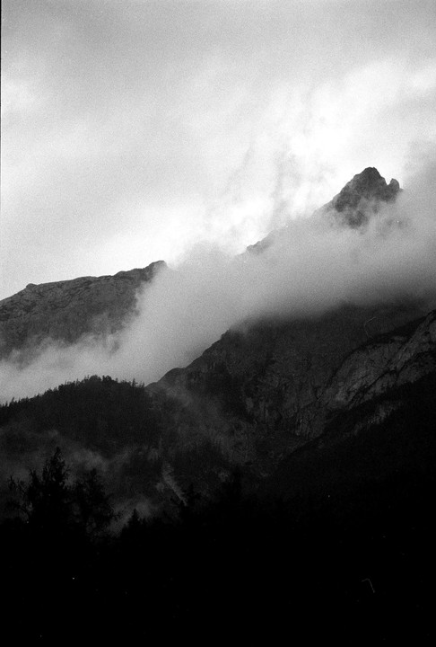 Clouds obscure the peaks of a mountain range with a dark forest in the foreground.