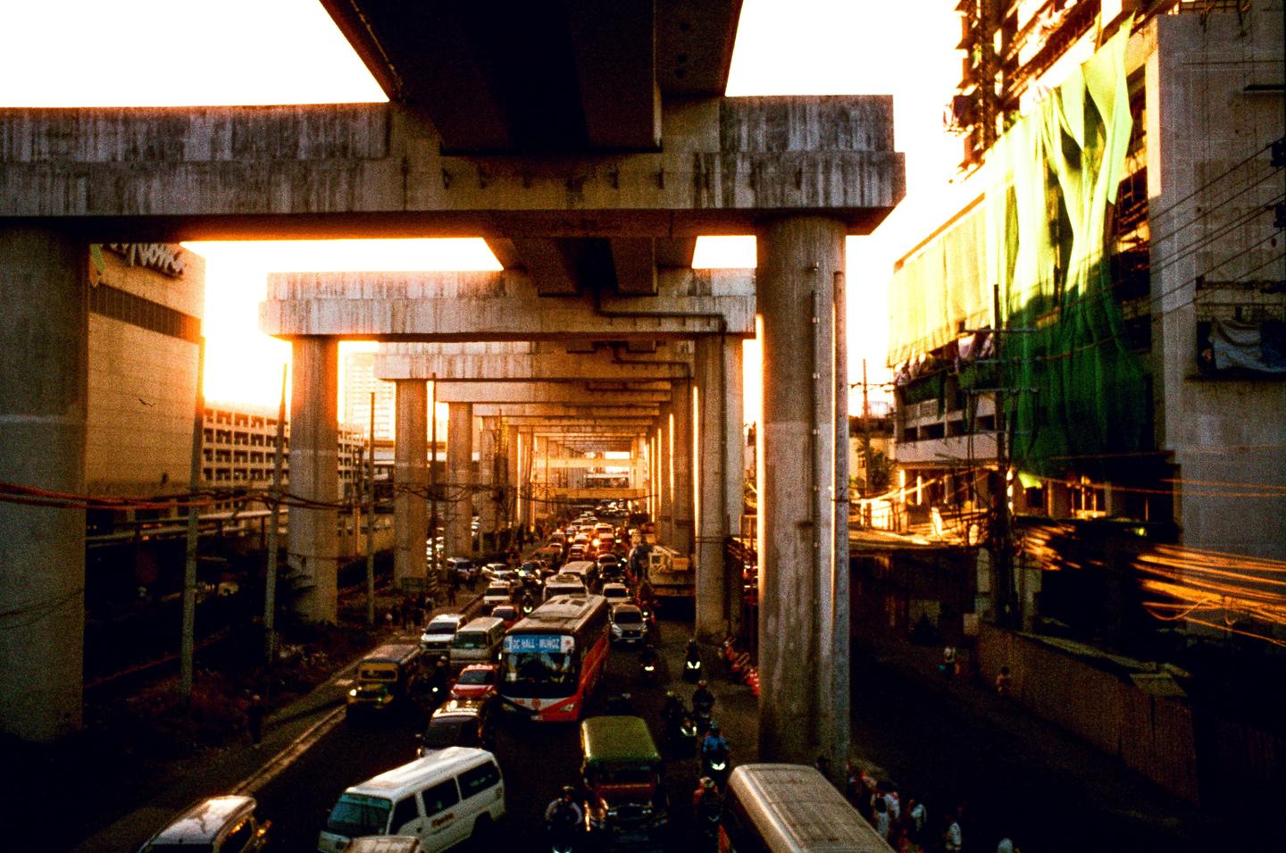 A busy street with heavy traffic is visible under a series of concrete overpasses during golden hour.