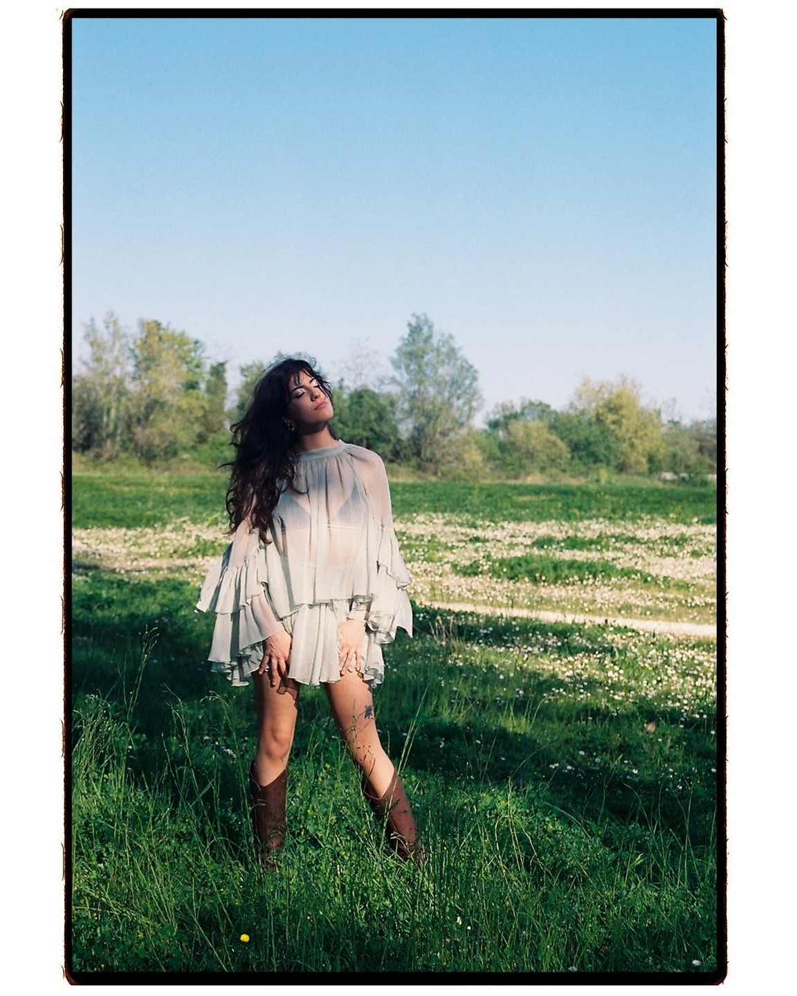 A woman stands in a field of tall grass and small white flowers, looking up at the sky.