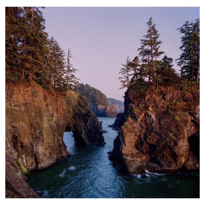 A body of water flows between two rocky cliffs covered in trees, with a natural arch visible on the left cliff.