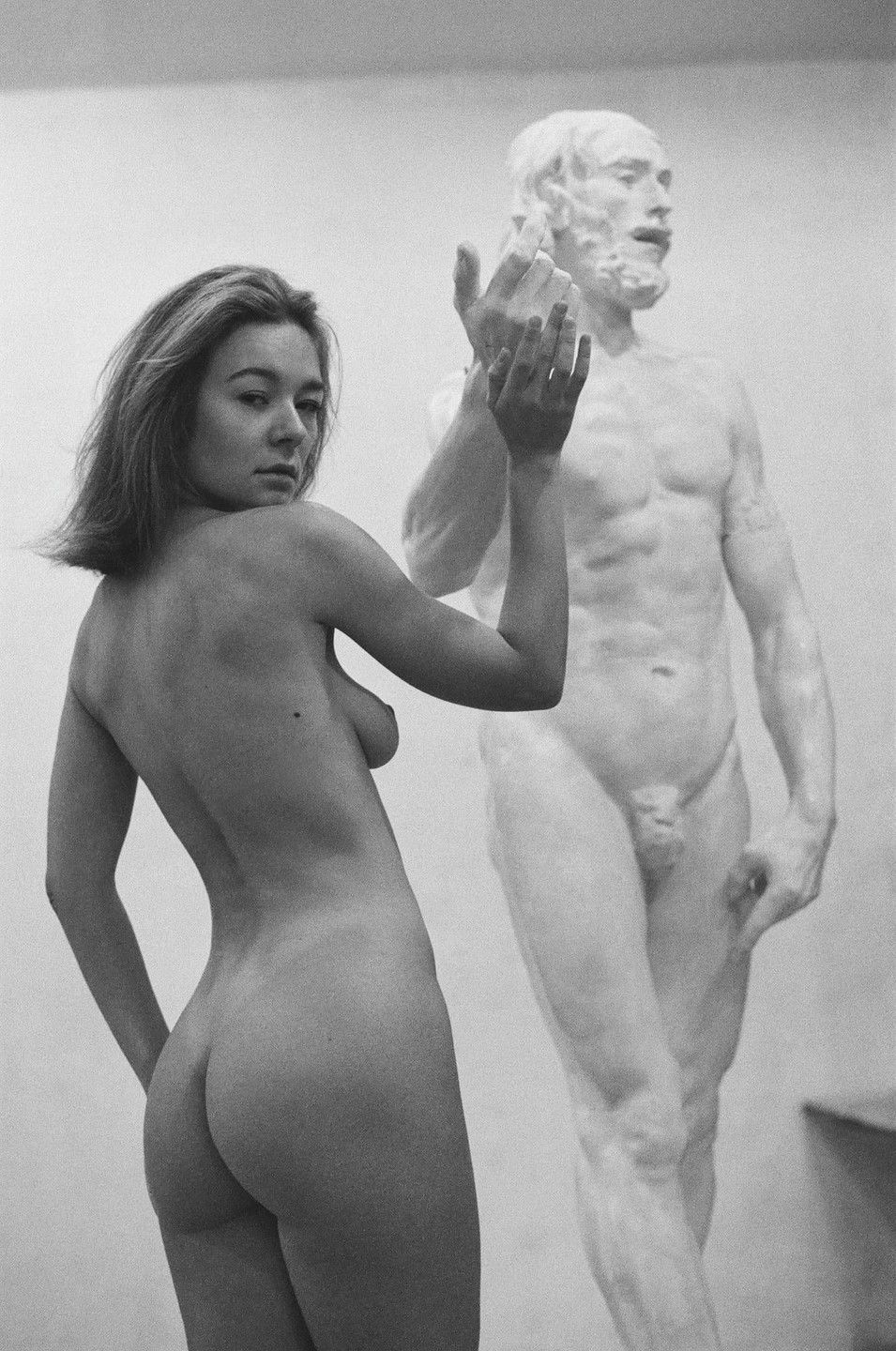 A nude woman stands next to a nude male statue in a studio.