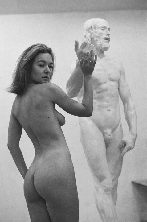 A nude woman stands next to a nude male statue in a studio.