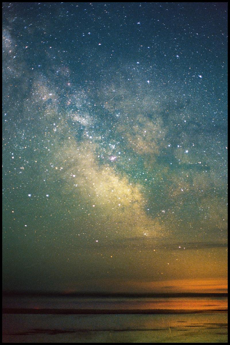 The milky way is visible above a body of water with a bright orange glow on the horizon.