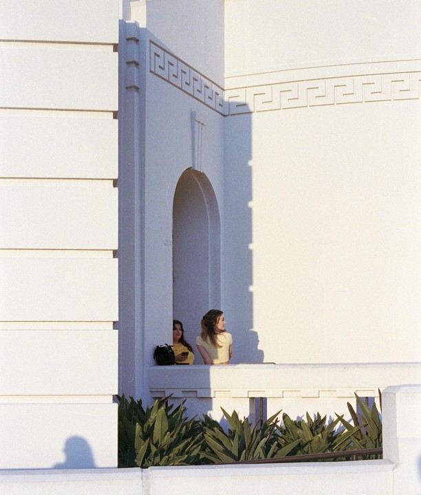 Two women stand on a white balcony with green plants below, one looking to the right and the other looking forward.