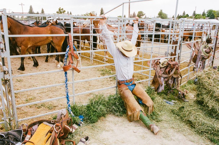 A man in a cowboy hat and chaps leans against a metal fence with horses in pens behind him.