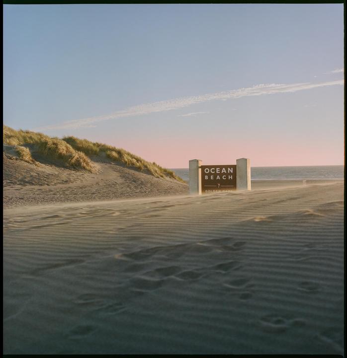 A sign for Ocean Beach stands on a sandy beach with dunes and the ocean in the background.