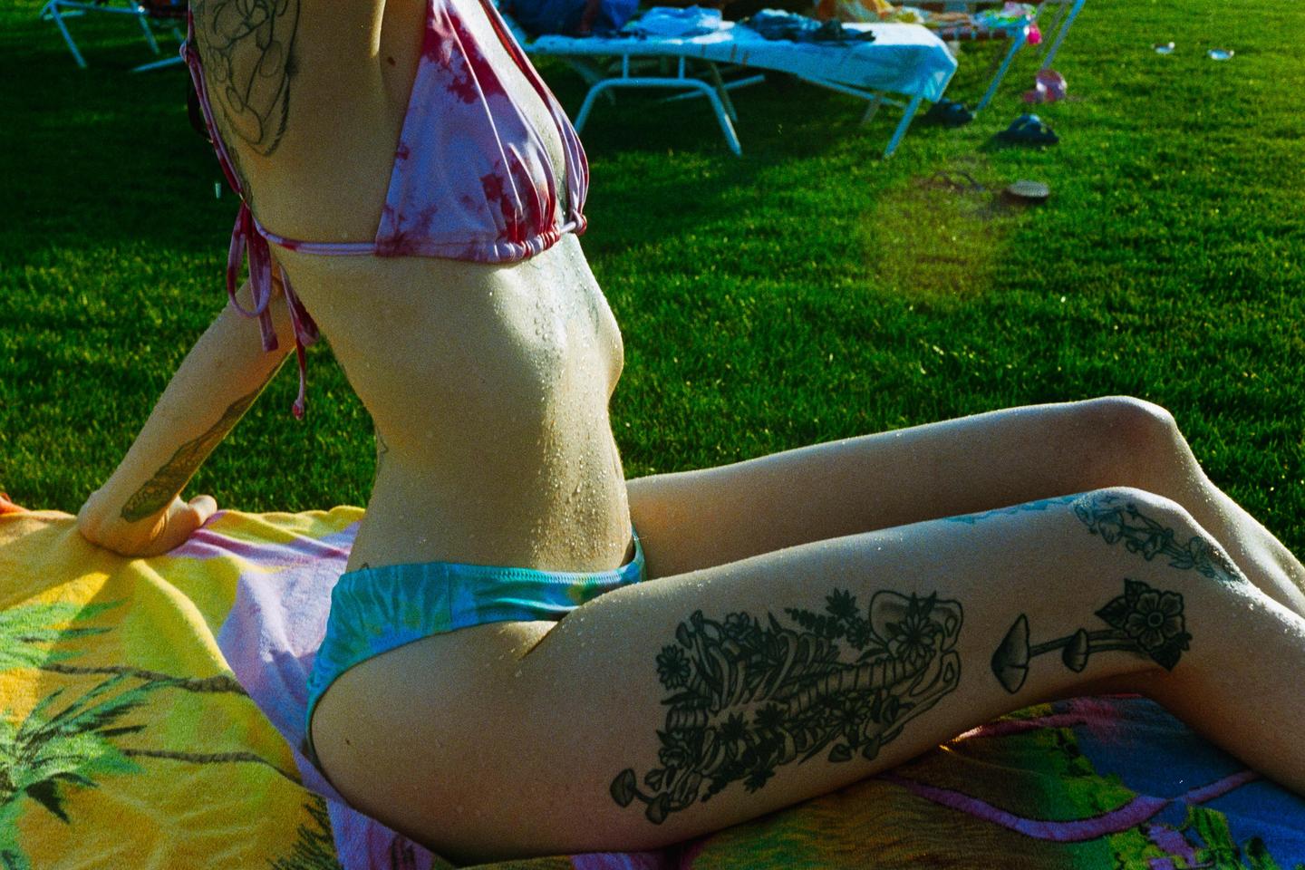 A woman with tattoos on her arm and leg sits on a towel in a grassy area.