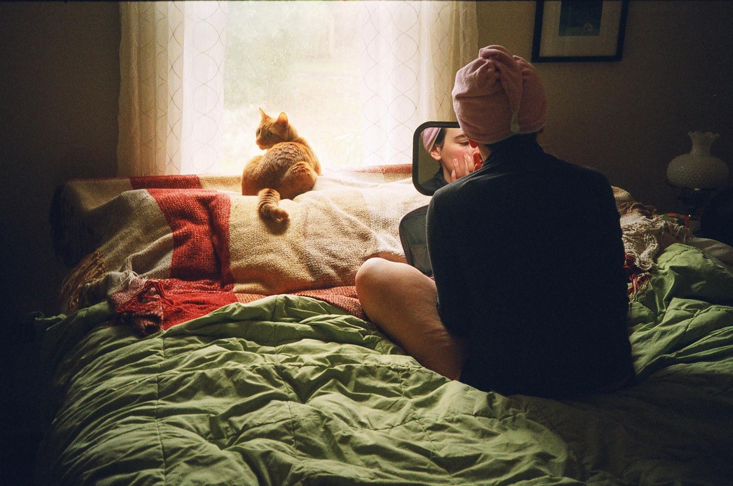 A woman with a towel on her head sits on a bed looking into a mirror while an orange cat sits on a pillow by a window.