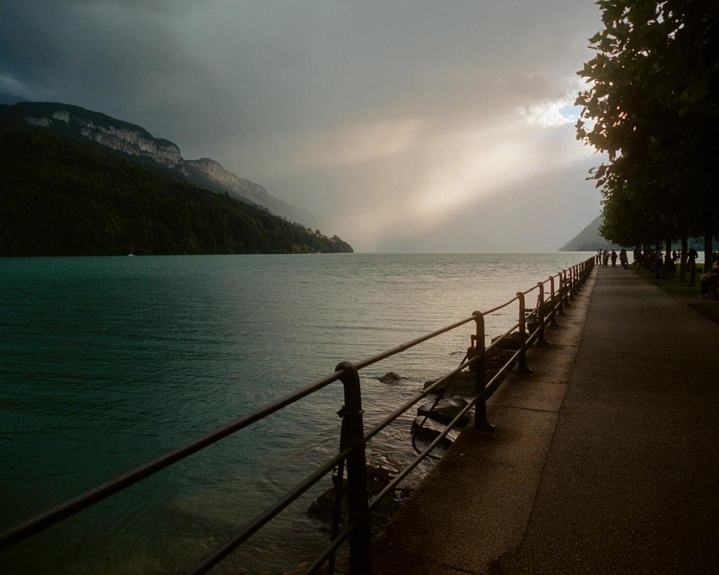 A lake with mountains and a paved path with a railing along the shore is illuminated by a ray of light from a cloudy sky.