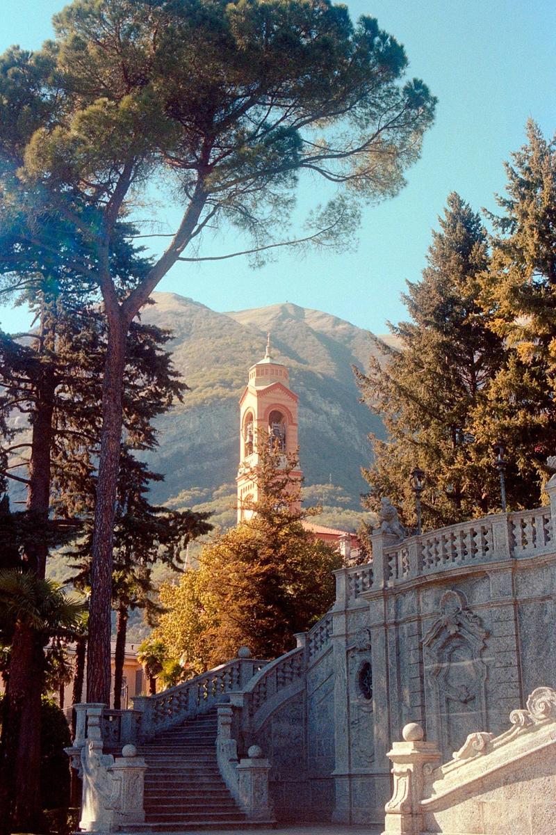 A church bell tower is visible behind a stone staircase and trees, with a mountain in the background.