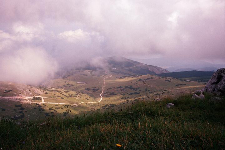 A winding road traverses a grassy mountain landscape under a cloudy sky.