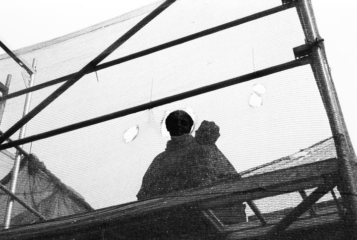 Two dark figures are visible through a mesh screen with structural beams.