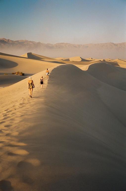 Three people walk along a sand dune in a desert landscape with mountains in the distance.