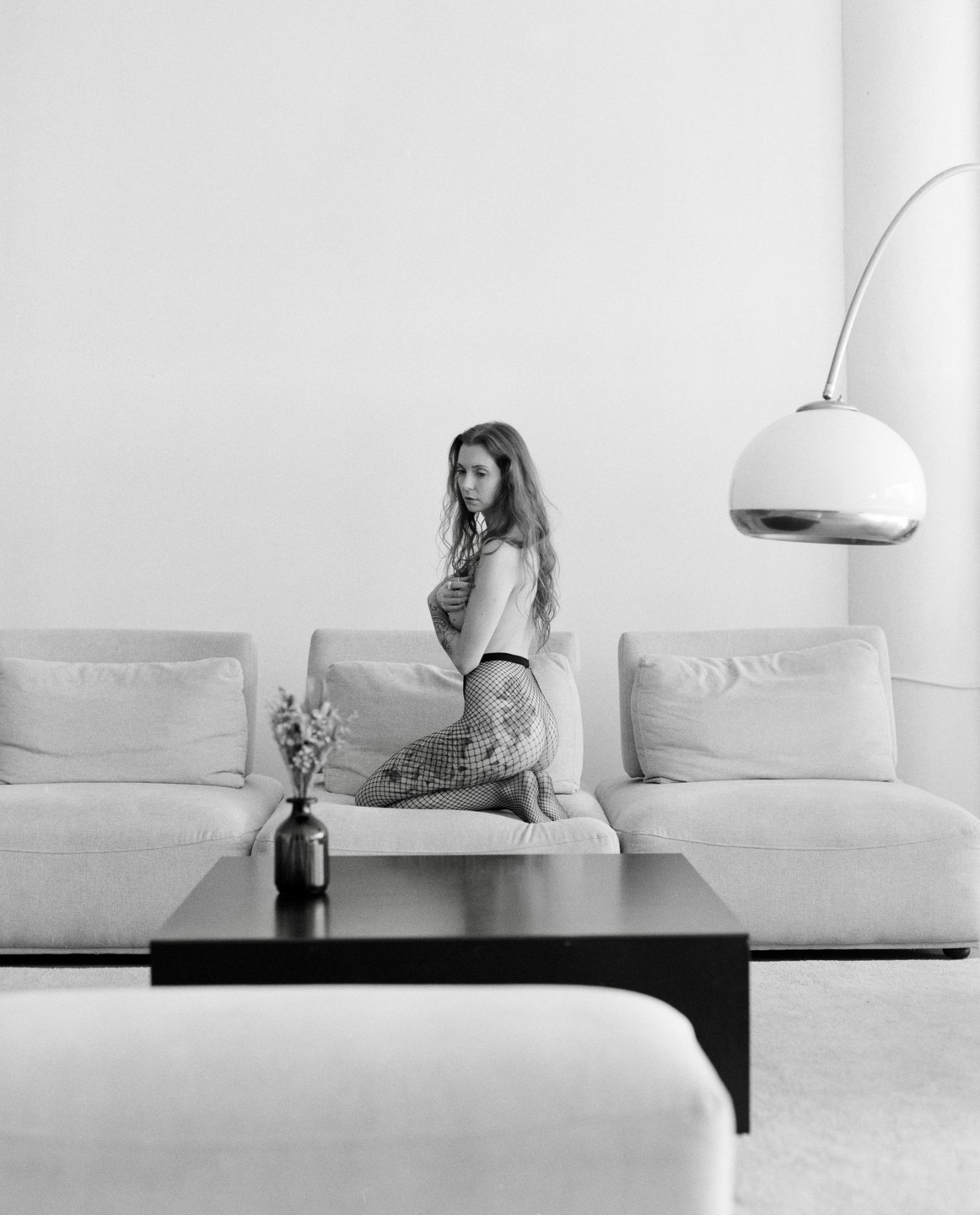 A woman kneels on a sofa in a living room, covering her chest with her arms.